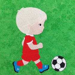 little boy with blond hair and a red football strip with blue boots kicking a football on a green background