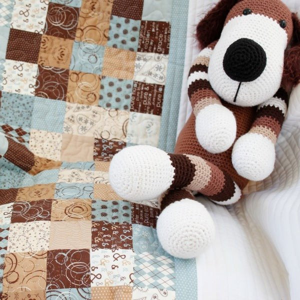 patchwork quilt with small squares of light and dark browns and blues with a white border. Sitting on the quilt is a brown toy dog.