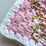 the corner of a crochet baby blanket, the corner is in white stitches and the other stitches are pink and green
